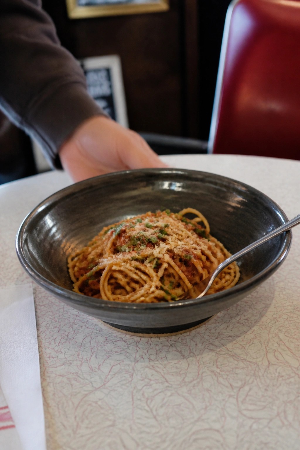 Brown-sleeved hand reaching toward dark bowl of spaghetti with fork