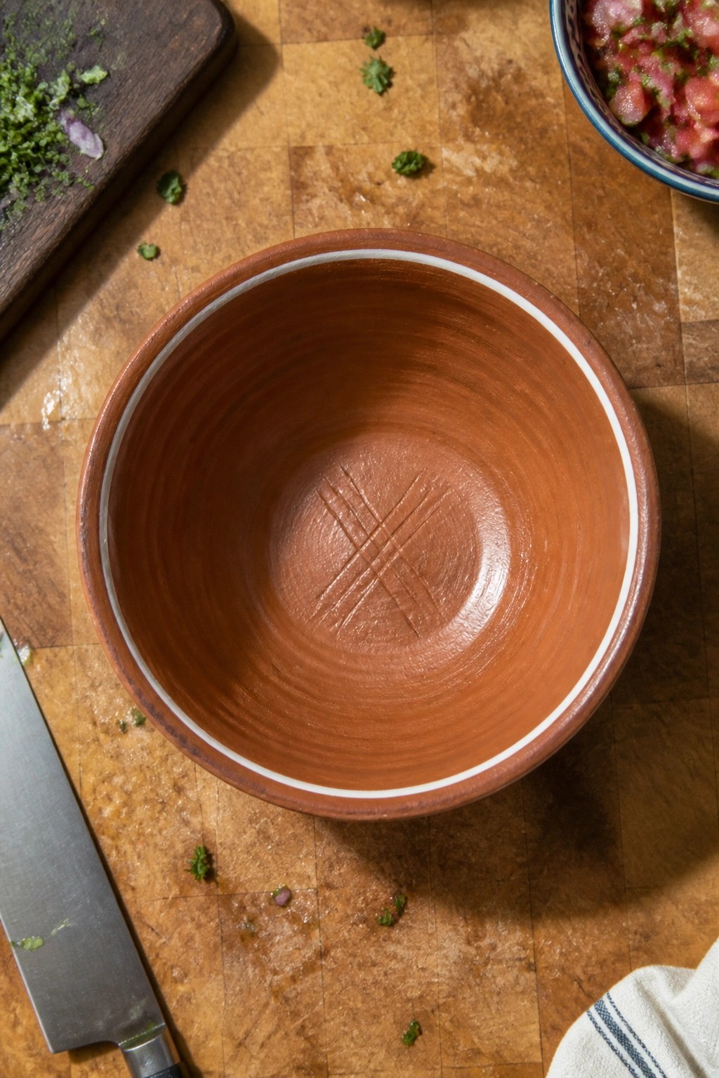 Empty brown terracotta bowl with white rim on wooden board amid herbs, knife, and ingredients
