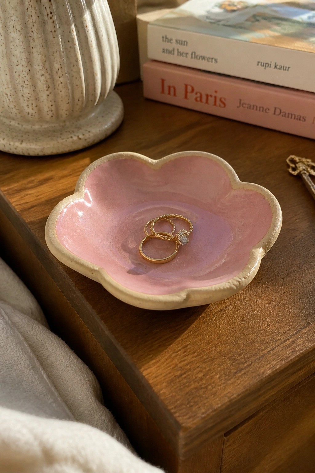 Pink flower-shaped ceramic dish holding gold rings on wooden table with books