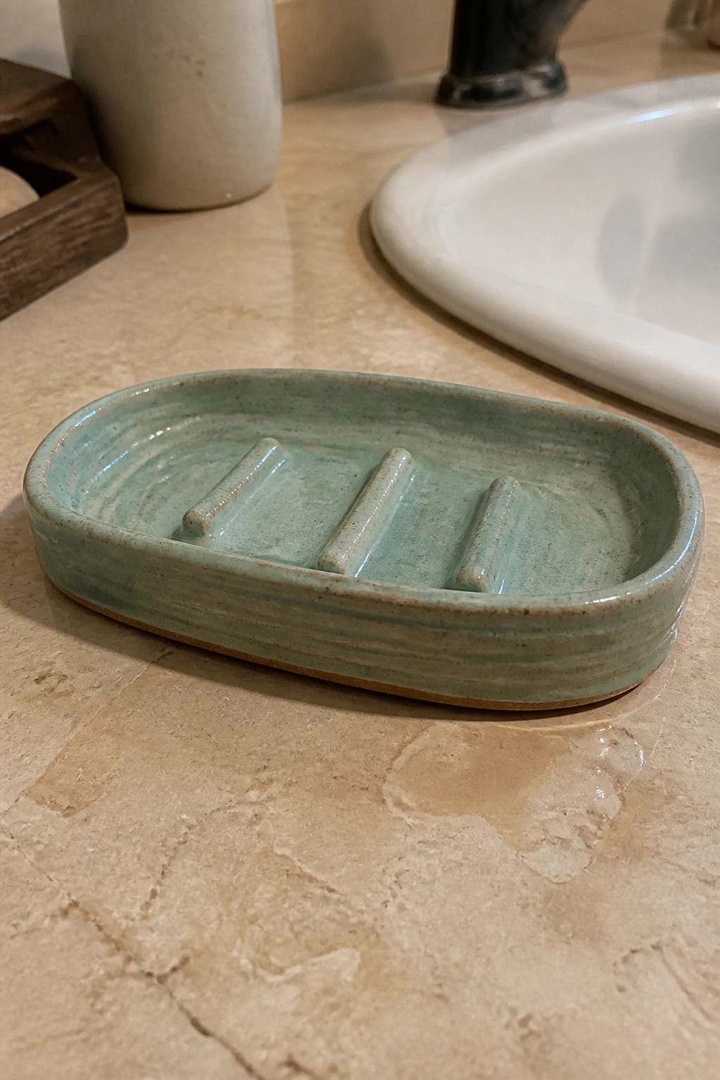 A light green oval ceramic soap dish with three raised ridges sits on a marble bathroom counter next to a white sink.