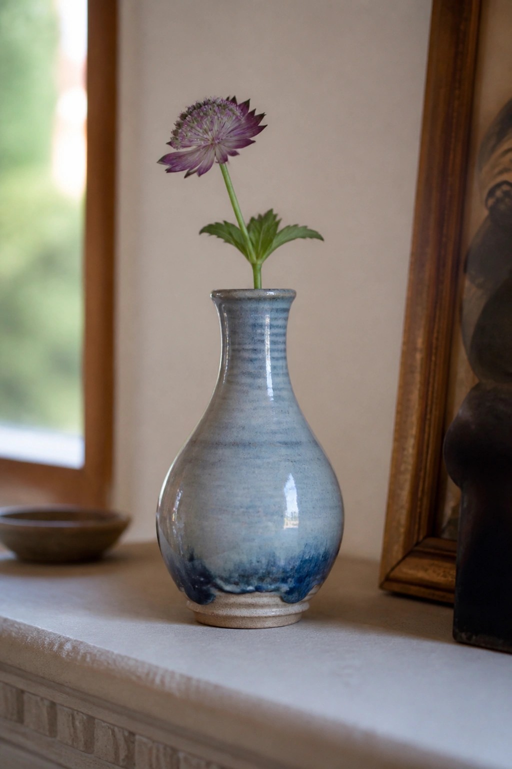 Blue ceramic vase with purple flower on sunlit windowsill beside frame
