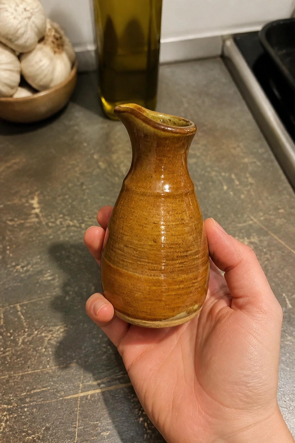 Hand holding small brown ceramic pitcher near garlic bowl and olive oil bottle