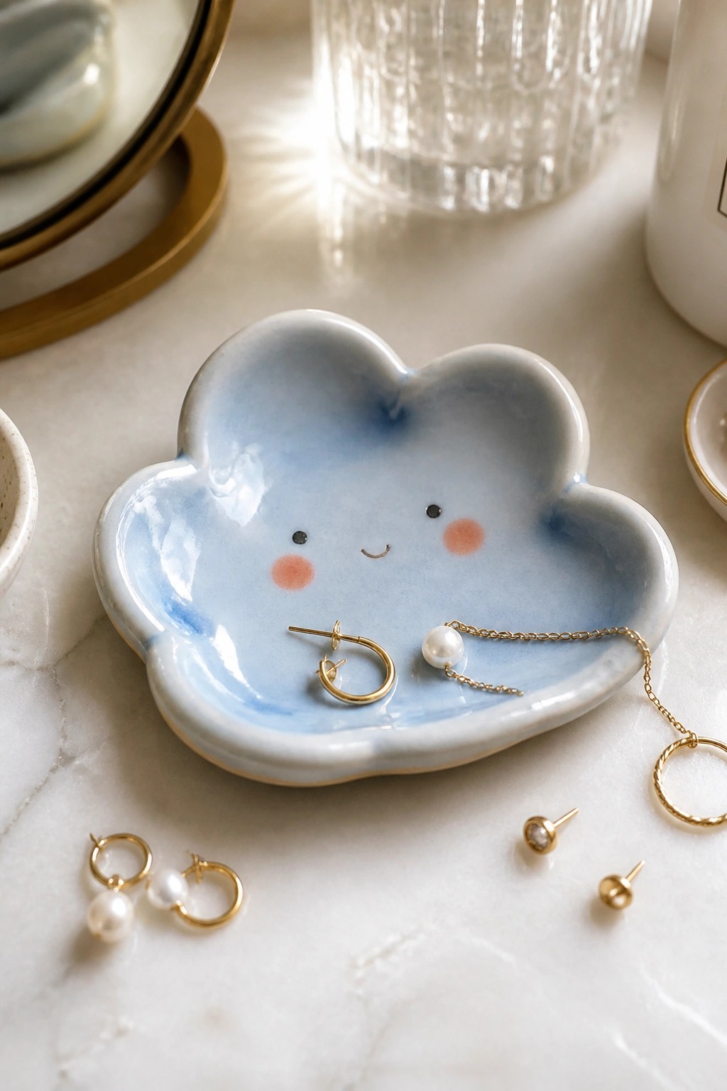 A blue glazed cloud-shaped ceramic dish with a painted face holds gold hoop earrings and a pearl necklace on a white marble surface.