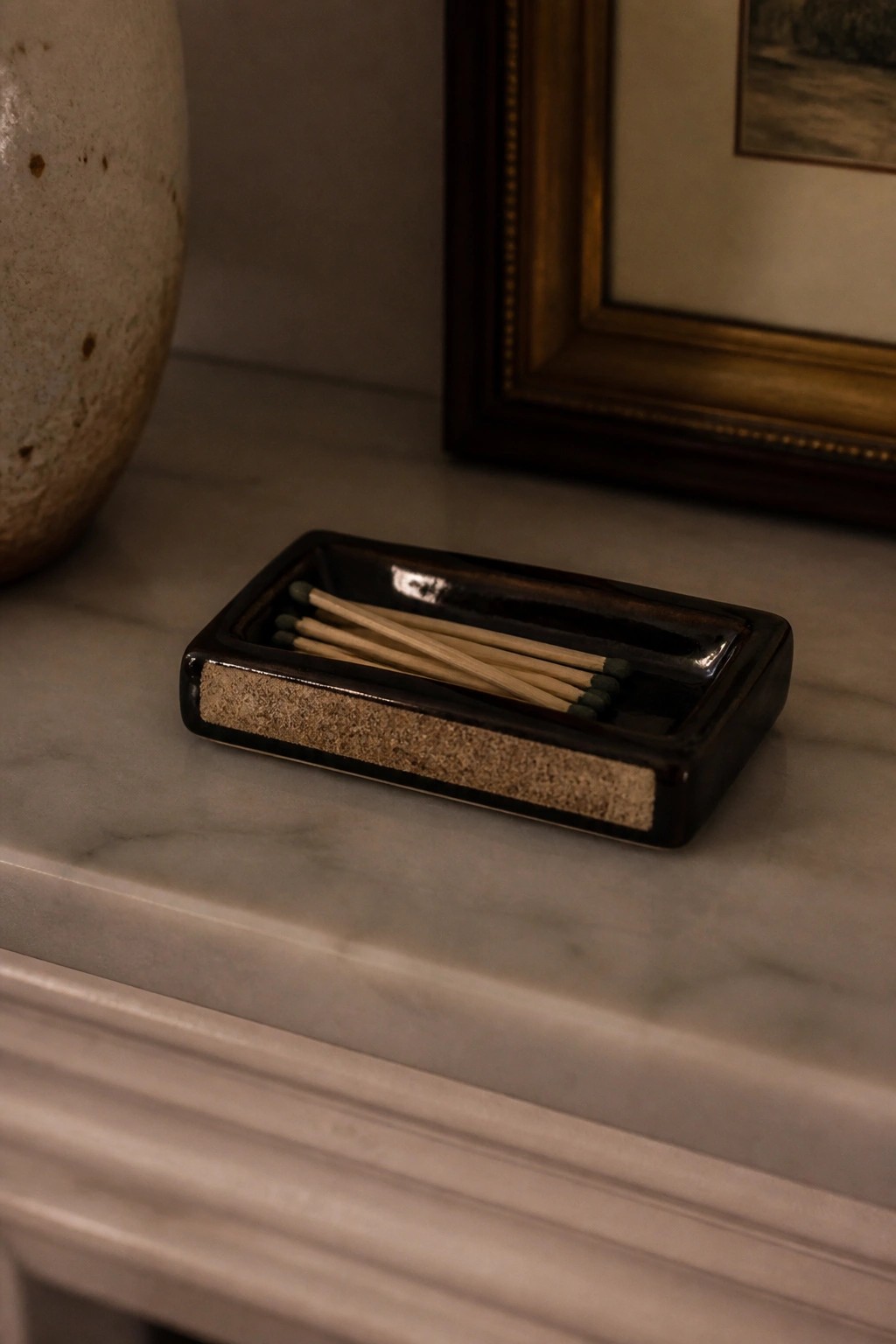 Dark ceramic match tray with wooden sticks on marble beside vase and frame.