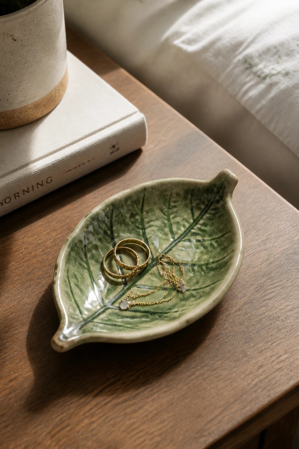 Green leaf-shaped ceramic dish holding gold rings and necklace beside book on wood table
