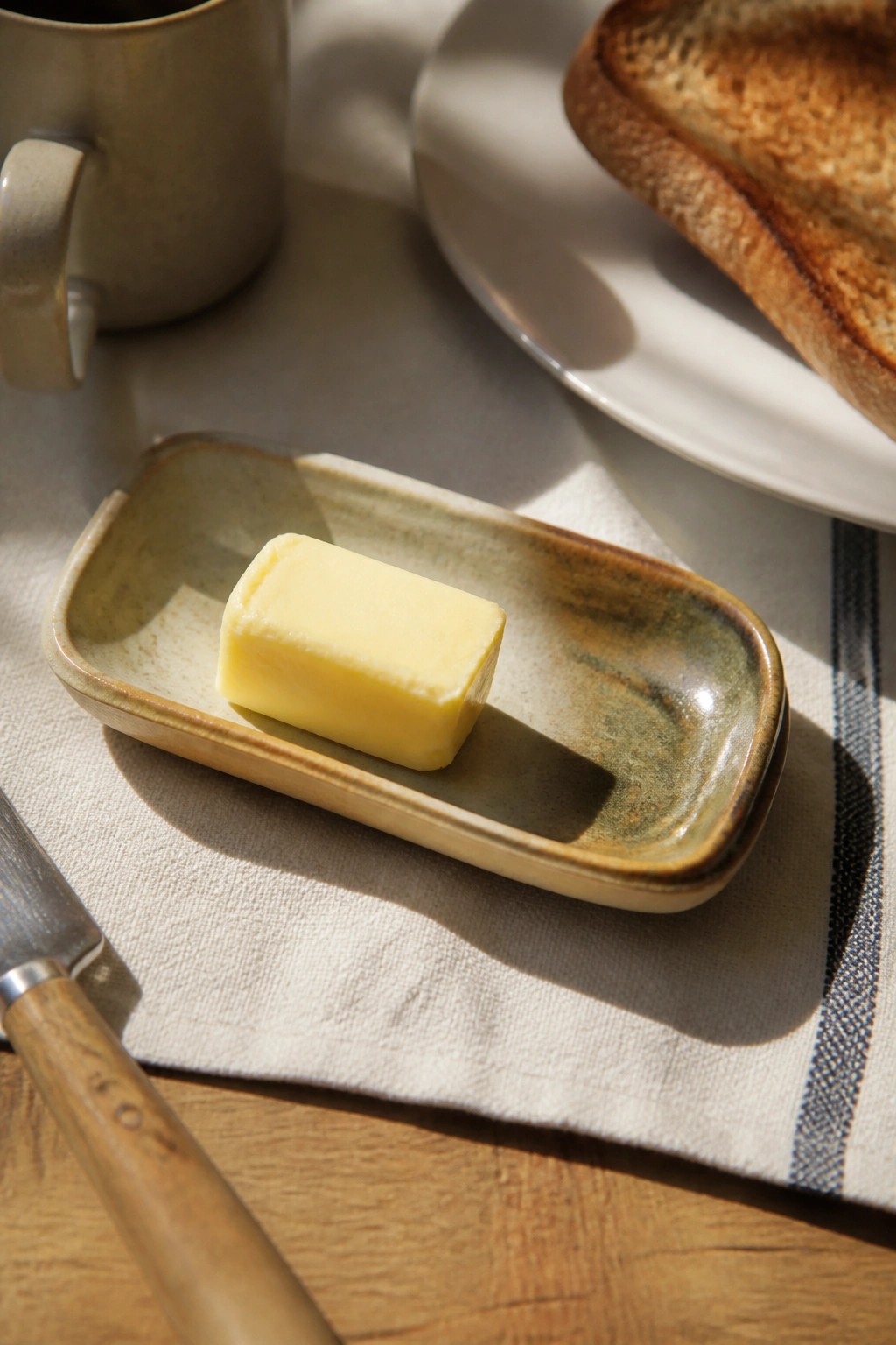 Butter on a small rectangular ceramic dish beside a knife on a tablecloth