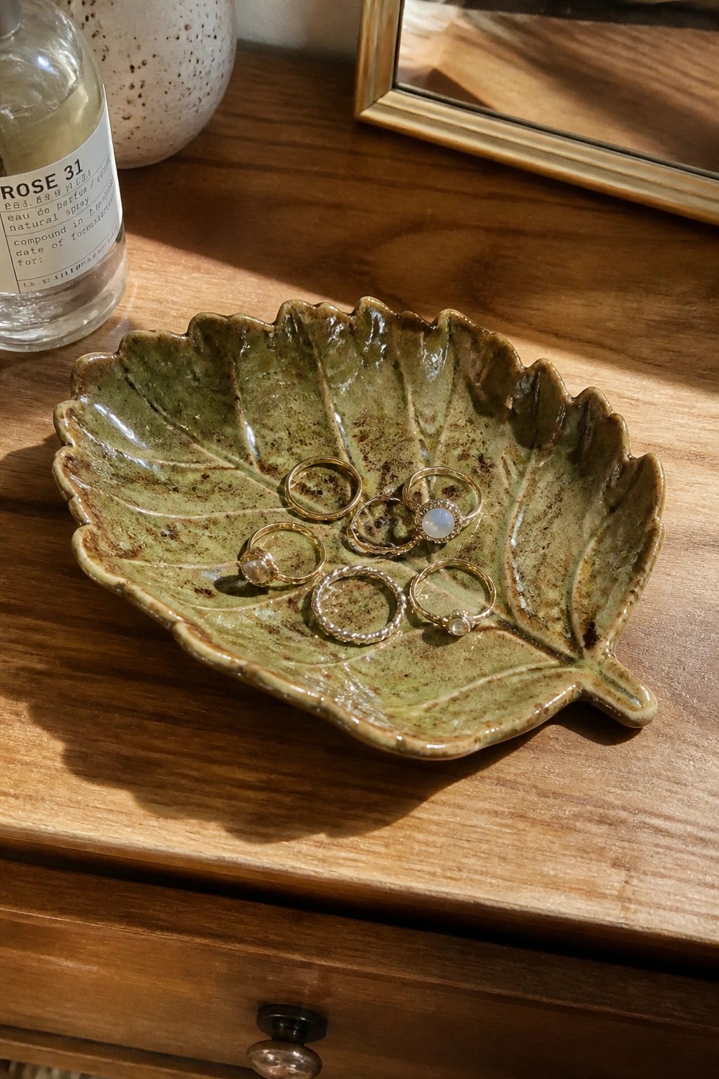 Green leaf ceramic dish holding gold rings beside perfume on wooden dresser