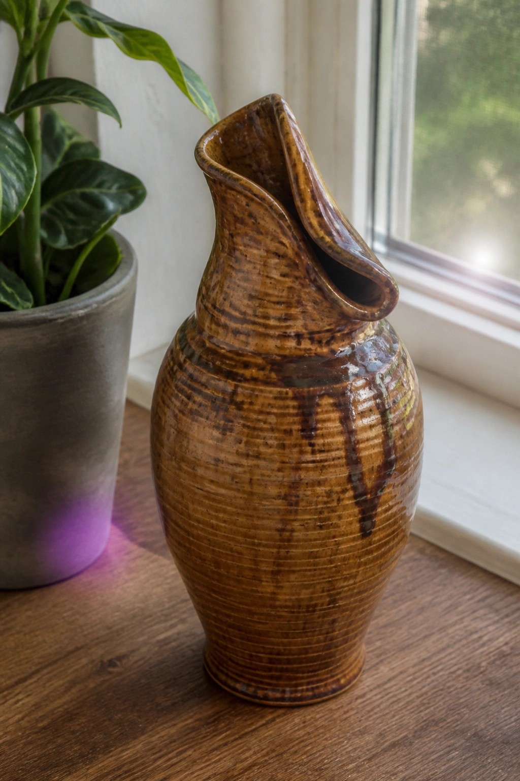 Brown ceramic vase with drippy glaze beside potted plant on wooden windowsill