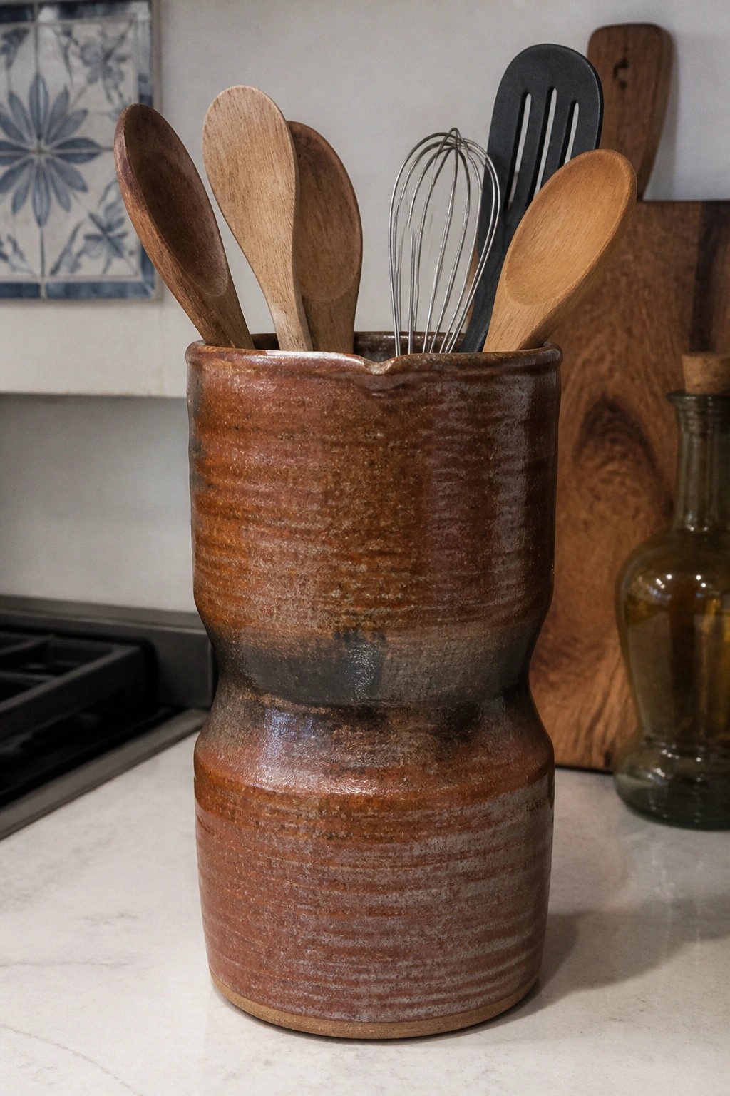 Rustic brown ceramic crock holding wooden spoons, whisk, and spatula on counter