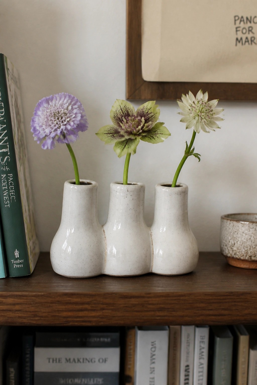 Three small white speckled ceramic bud vases joined together hold purple, green, and white flowers on a wooden shelf.