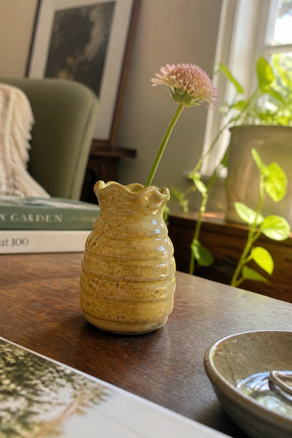Yellow ceramic vase with pink flower on wooden table beside books and bowl