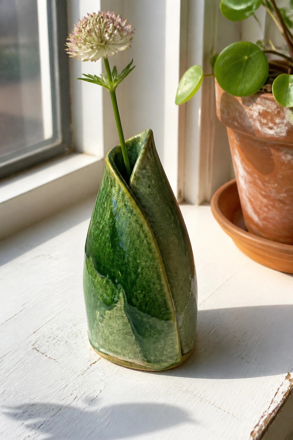 Green glazed ceramic vase with white-pink flower on sunny windowsill