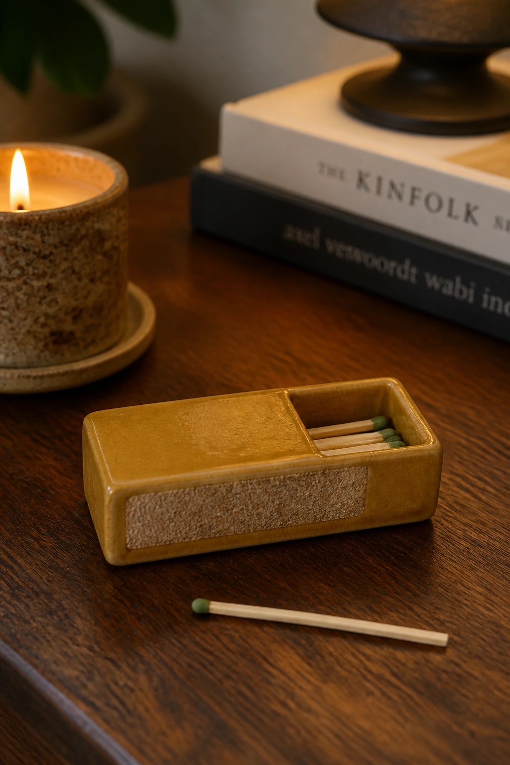A small rectangular mustard-yellow ceramic matchbox with matches inside sits on a wooden table beside a lit candle and stacked books.