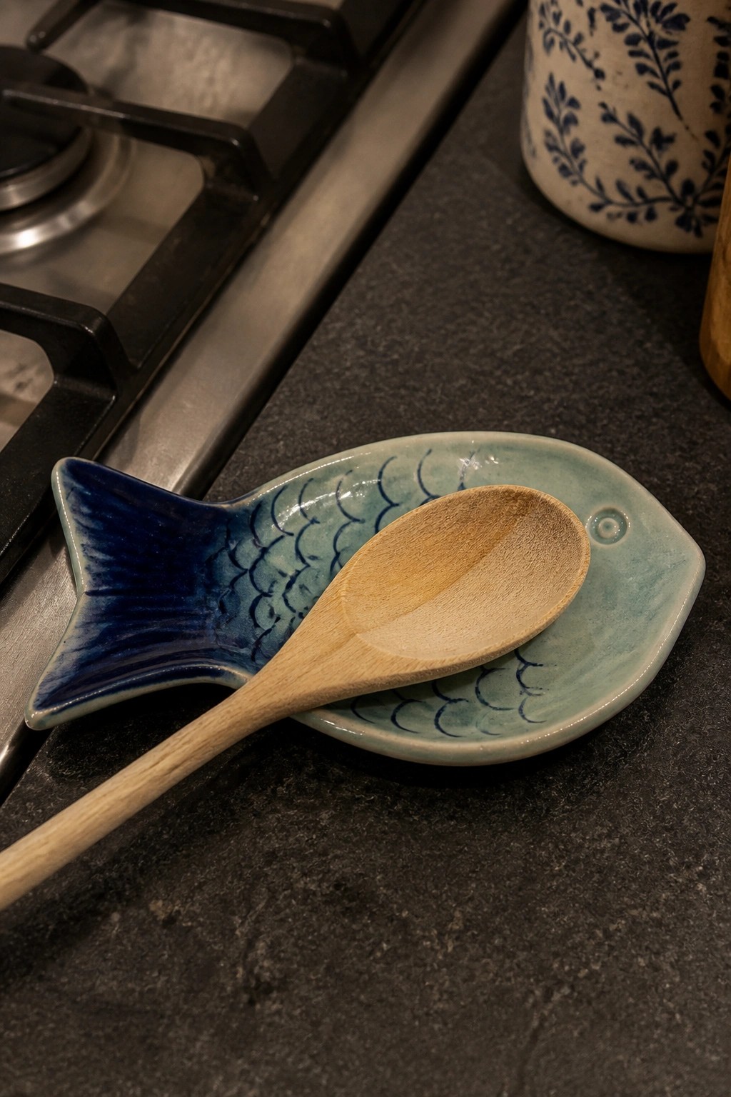 Wooden spoon on blue-green fish-shaped ceramic dish atop dark kitchen counter.