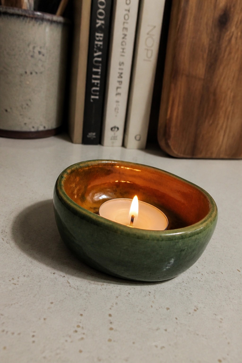 Green ceramic bowl with lit tea light candle on light surface near books
