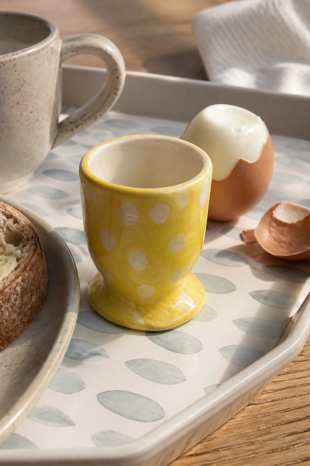 Yellow polka-dotted egg cup with boiled egg on patterned tray beside bread and mug.
