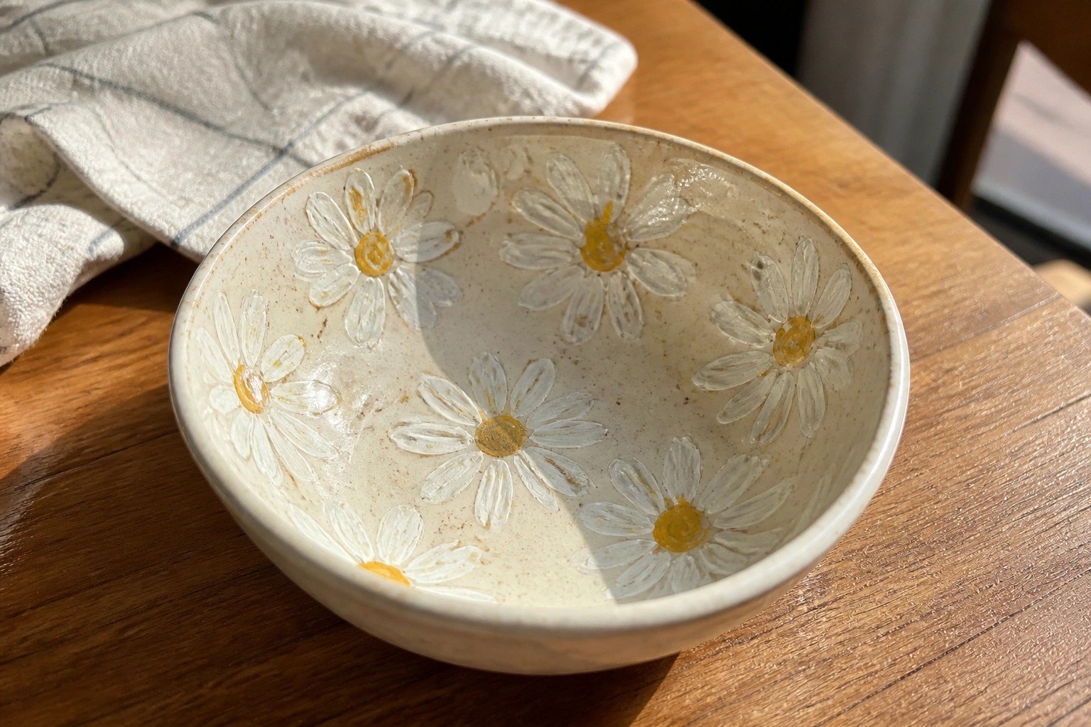 Ceramic bowl with blue-outlined daisies on wooden table beside checkered cloth.