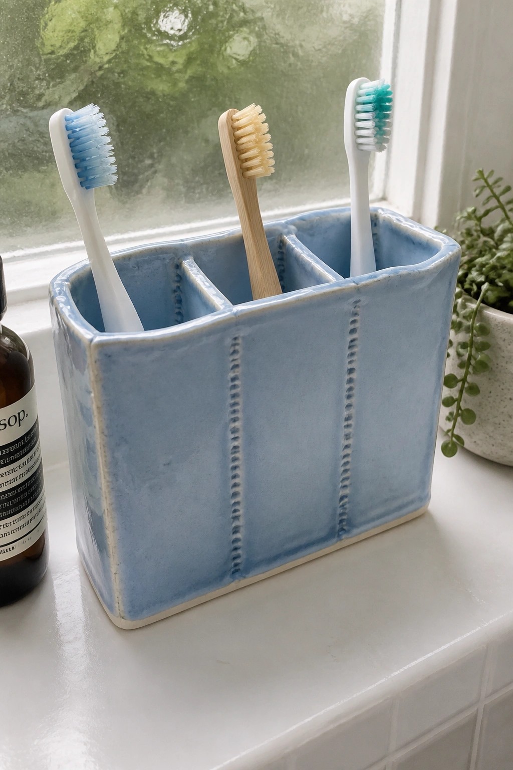 Blue ceramic holder with three toothbrushes on white windowsill beside plant