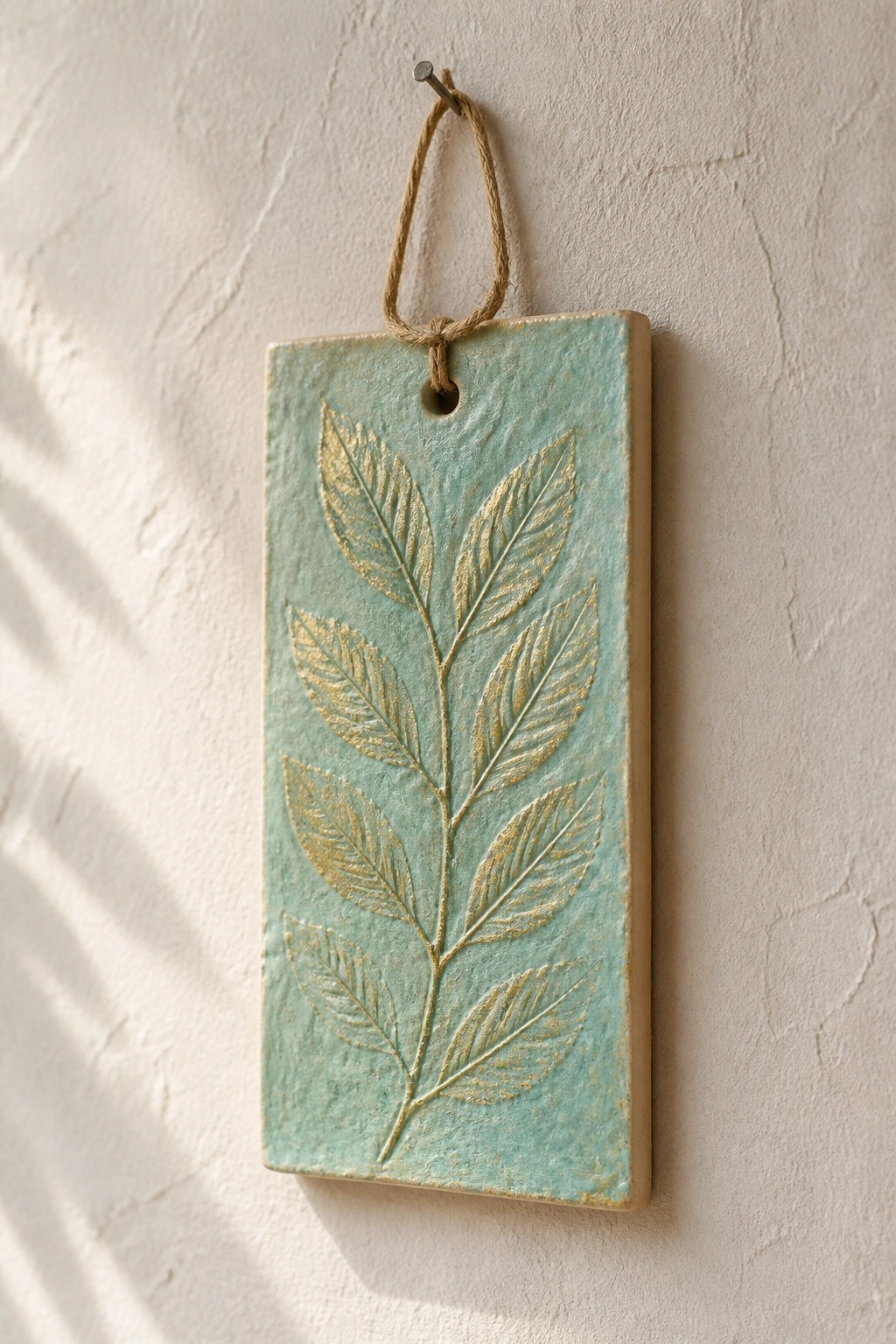 Turquoise ceramic plaque with gold-embossed leaves hanging from twine on white wall