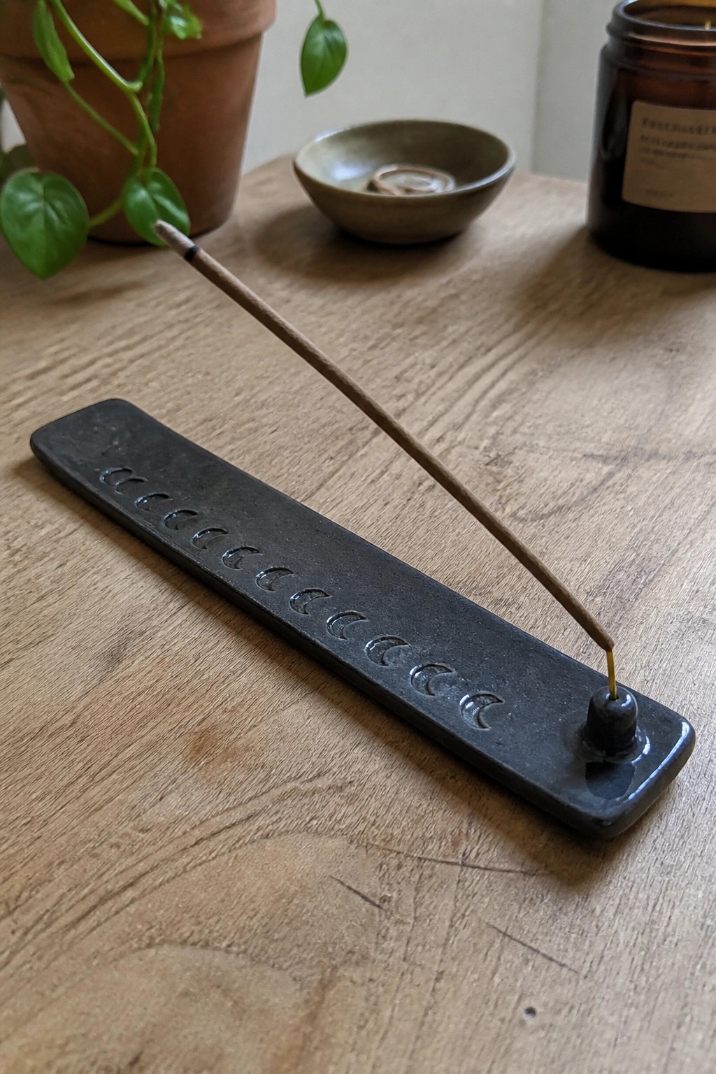 Burning incense on black moon phase holder atop wooden table with potted plant