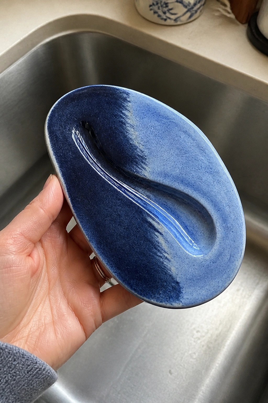 Hand holding oval blue ceramic dish with curved indentation over kitchen sink.