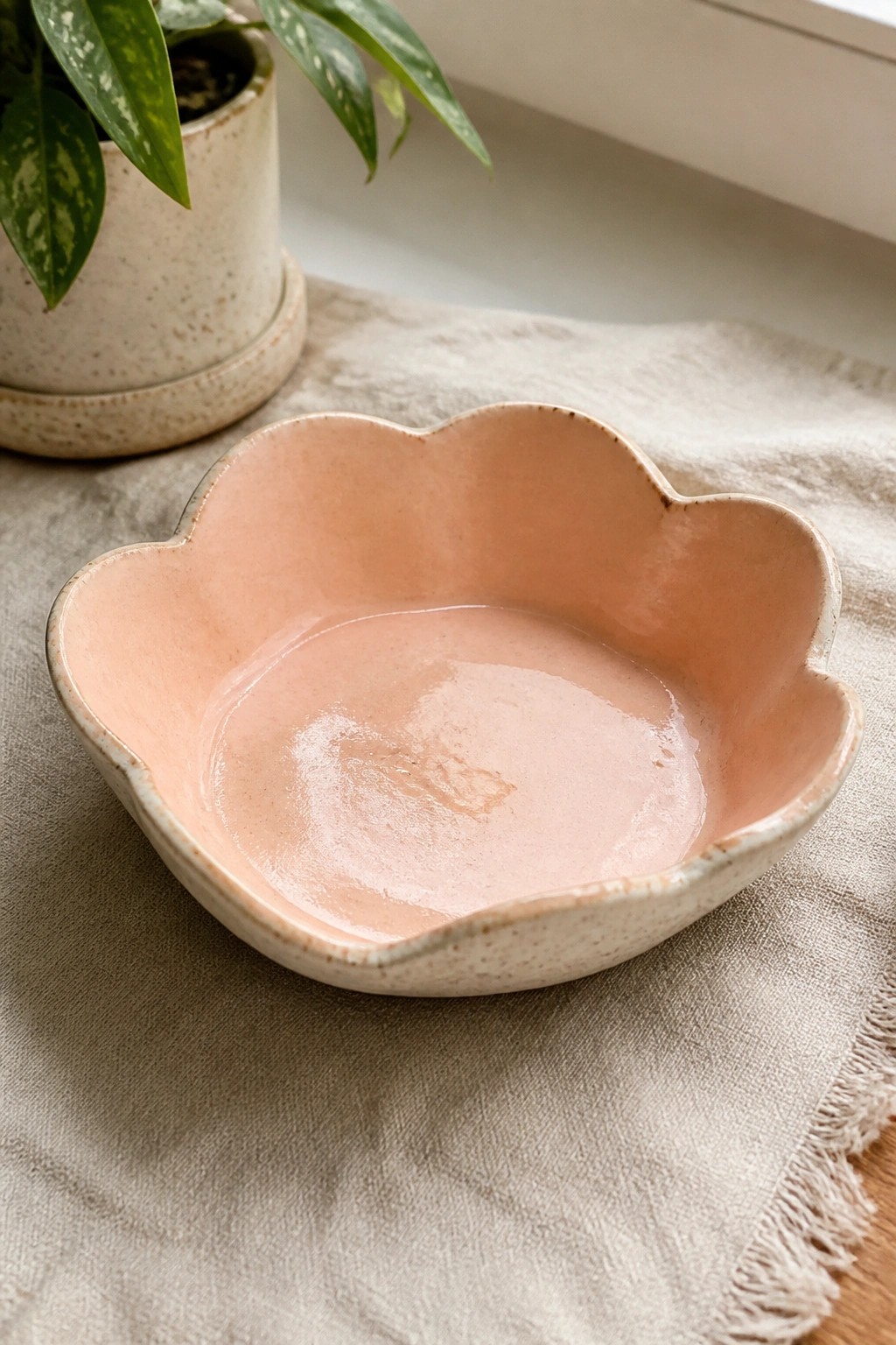 Pastel pink scalloped ceramic bowl on beige linen with potted plant nearby.