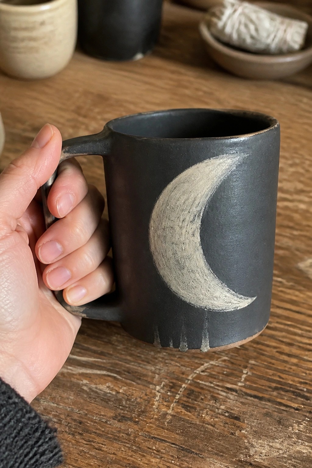 Hand in dark sweater holding black mug with white crescent moon on wooden table.
