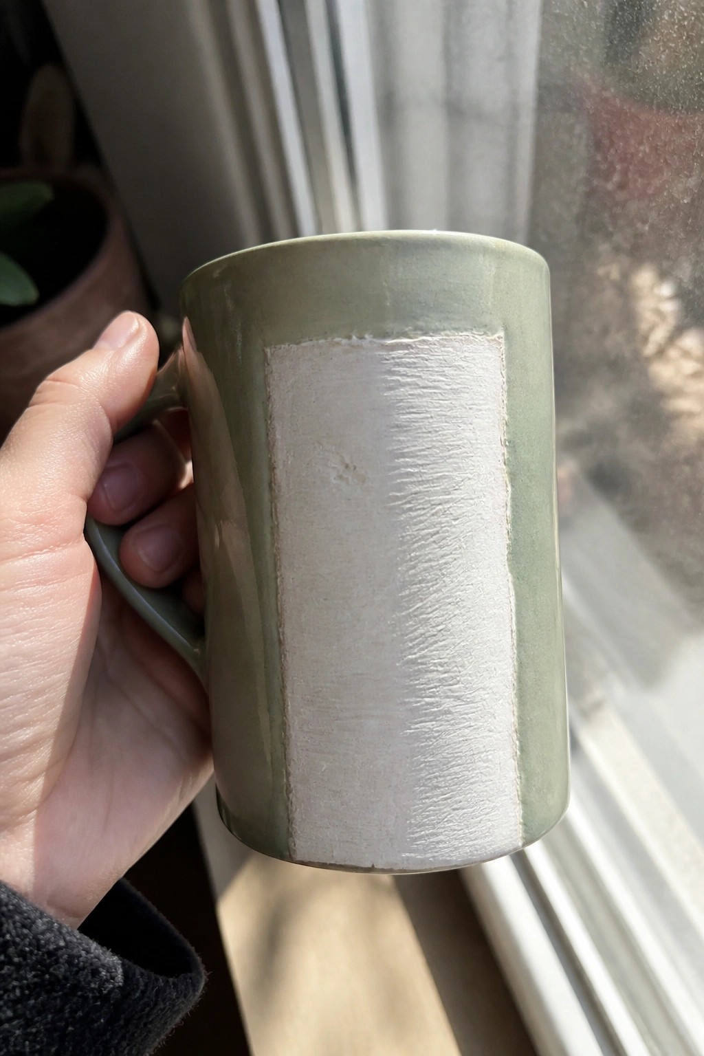 Hand holding sage-green ceramic mug with large white textured rectangular patch.