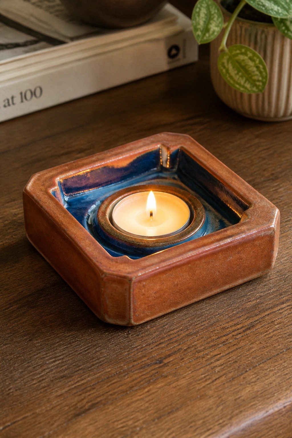 Lit tea light candle in square brown ceramic holder with blue glaze on wood