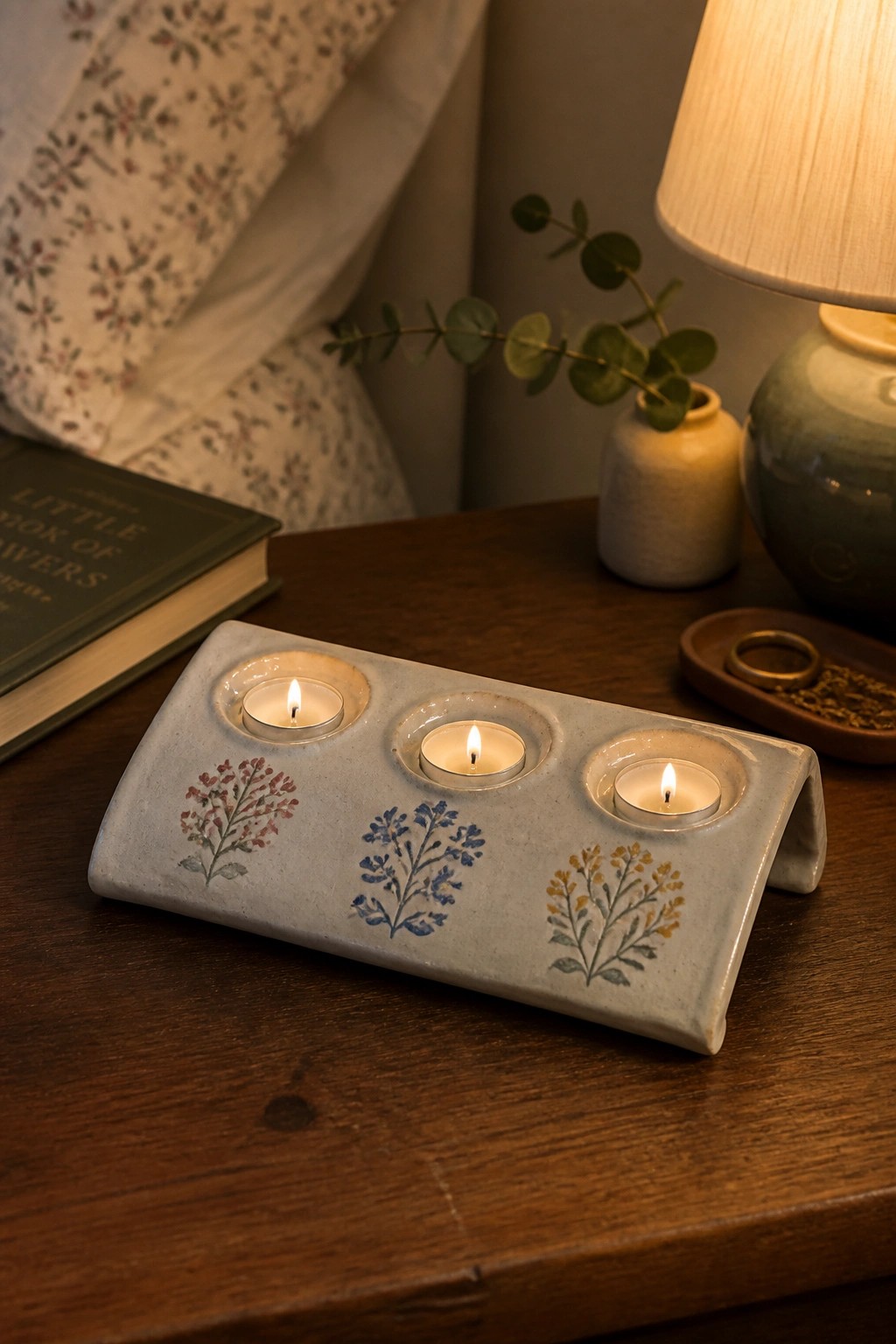 Three lit candles in floral ceramic holder on wooden table with book and lamp