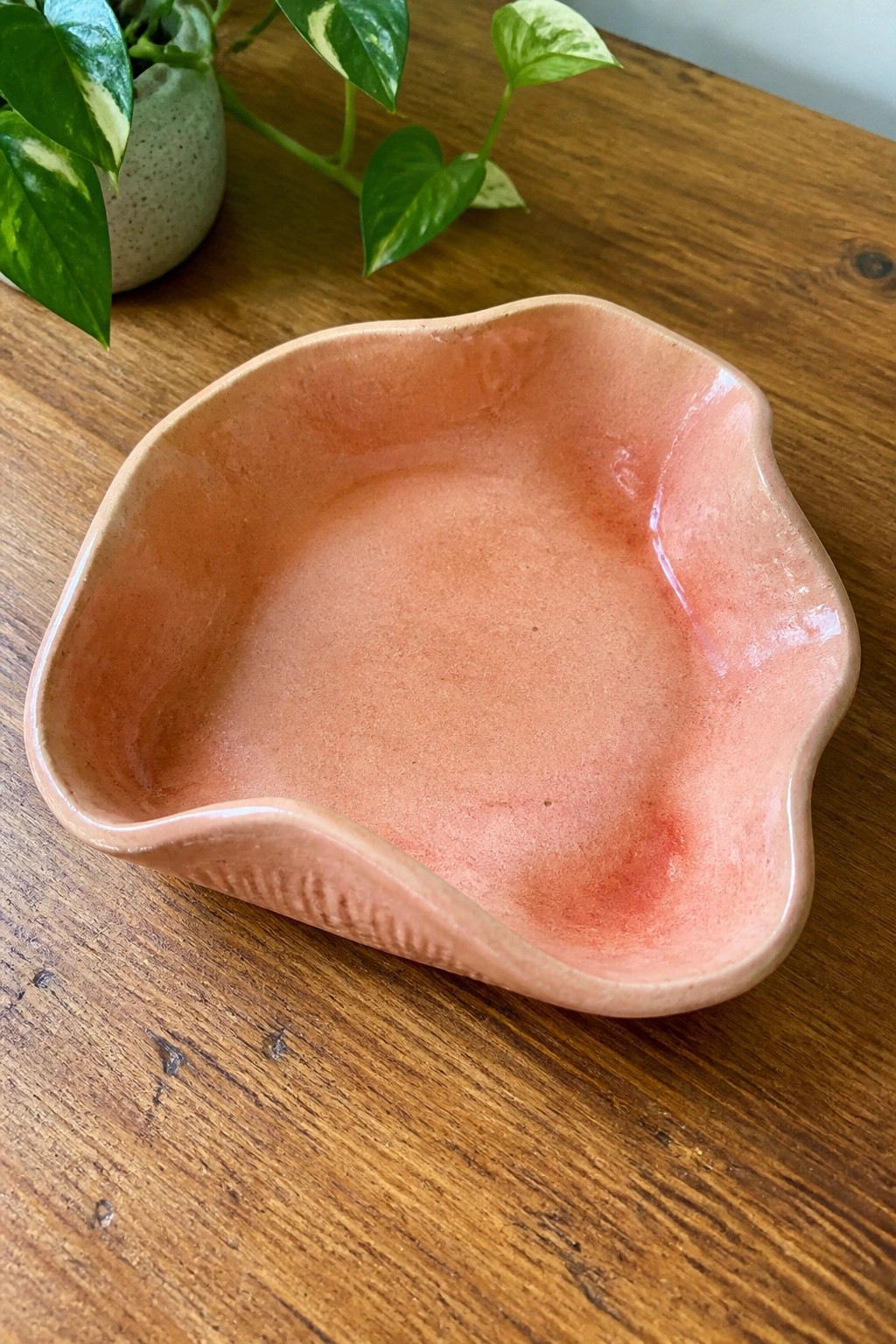 Irregular pink ceramic bowl with wavy rim on wooden table beside green plant