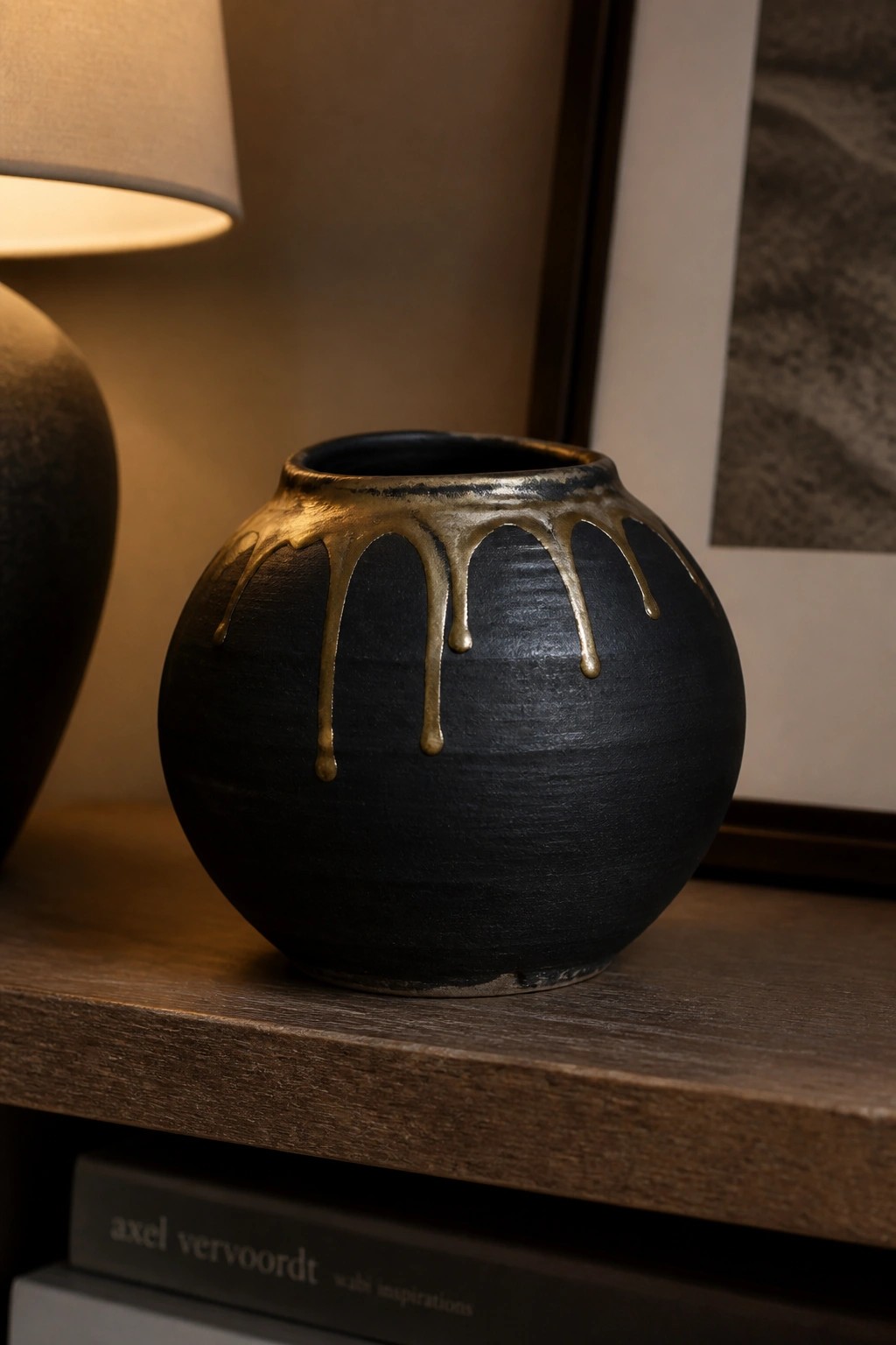 Black round vase with dripping gold glaze on wooden shelf with lamp nearby