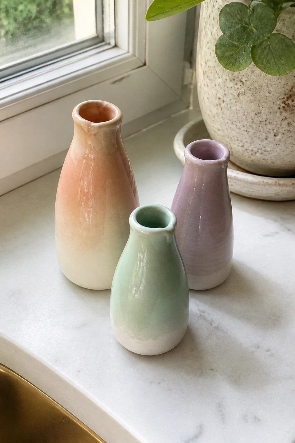 Three pastel ceramic vases on white marble near window with potted plant