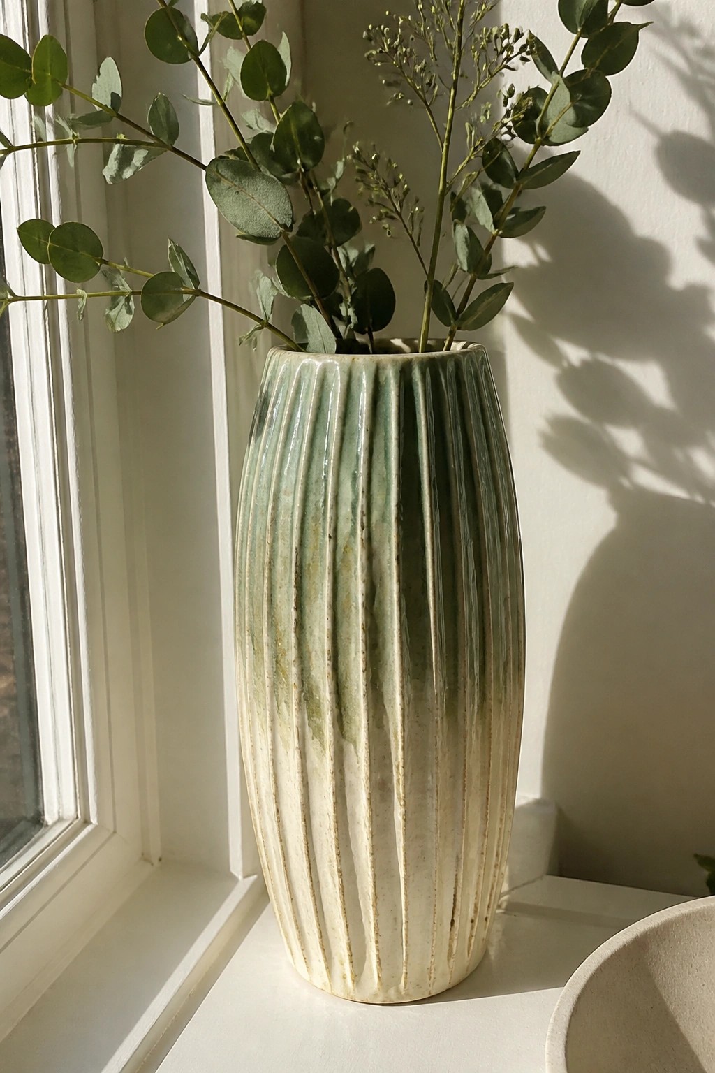 Ribbed ceramic vase with green glaze holding eucalyptus branches on sunlit windowsill