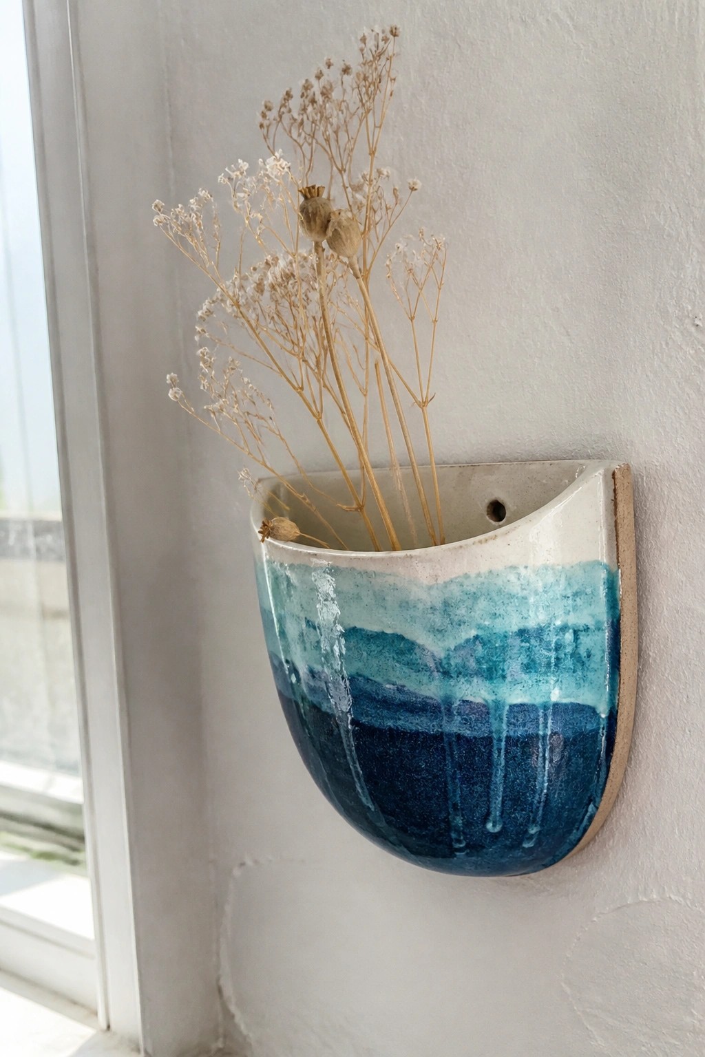 Blue ceramic wall vase with dripping glaze holding dried flowers on white wall