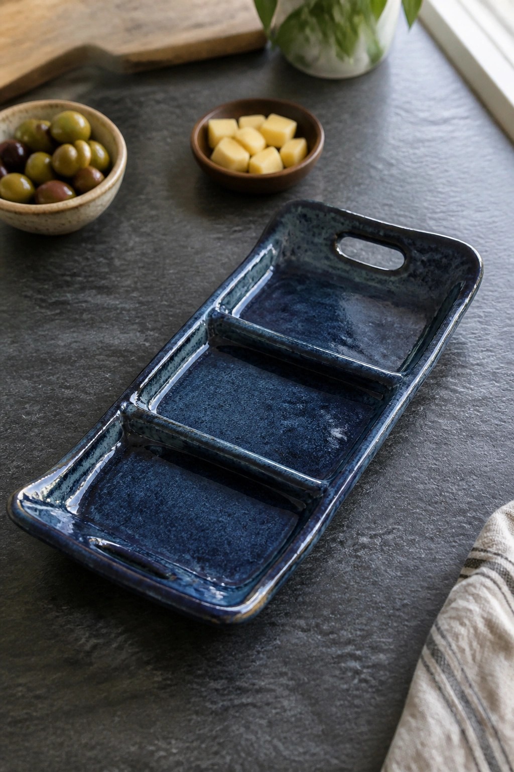 A blue ceramic divided serving tray with cutout handles on a dark countertop, next to bowls of olives and cheese.
