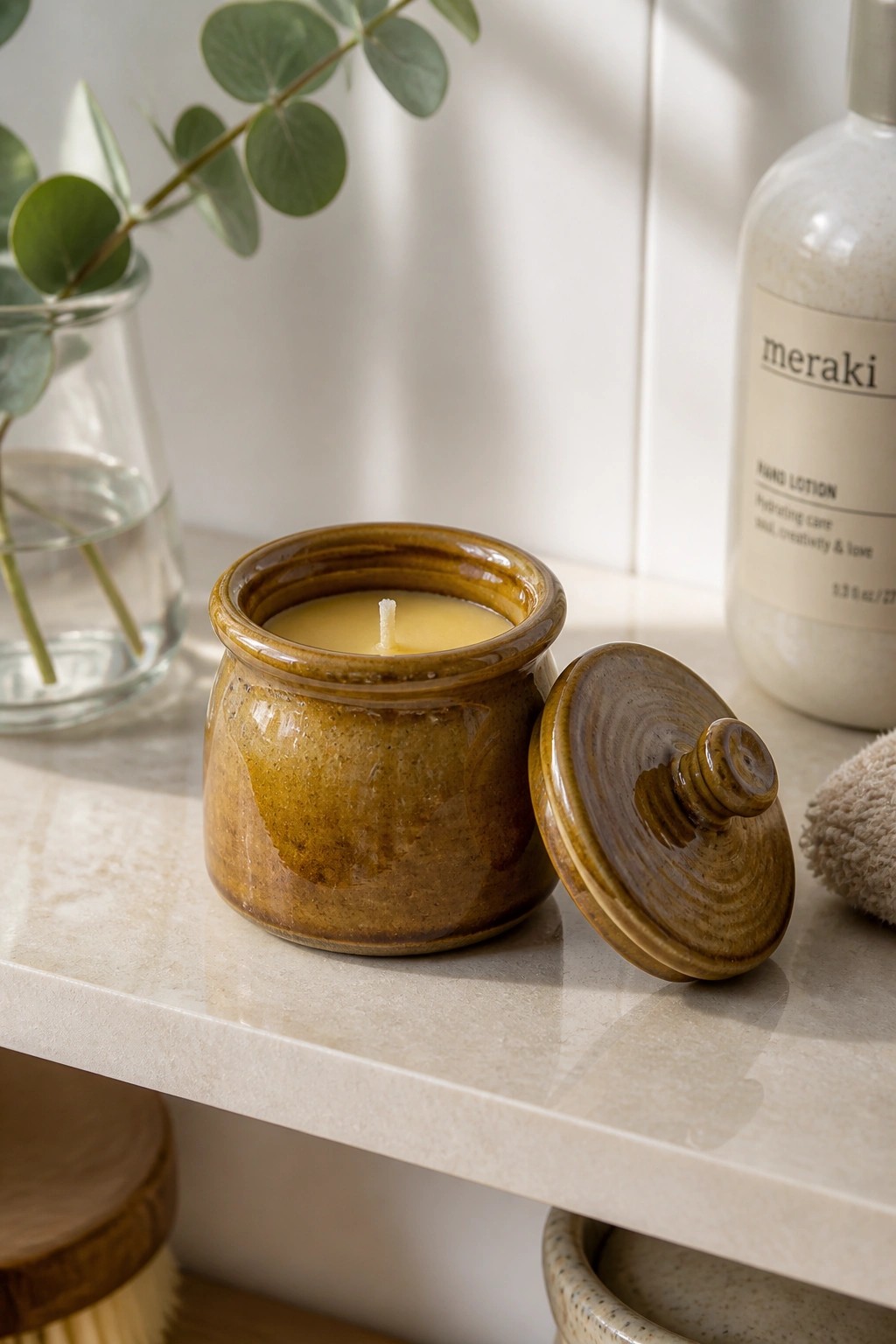 Open brown ceramic candle jar with lid on marble counter beside lotion and eucalyptus