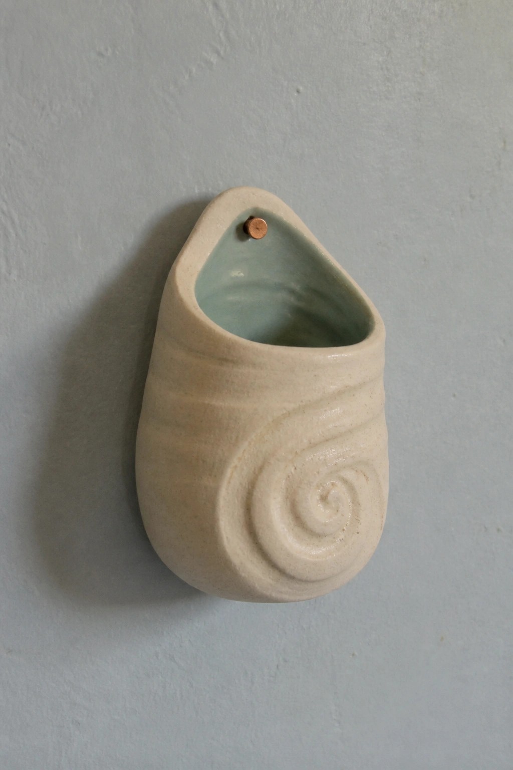 Beige ceramic spiral vase with turquoise interior mounted on gray wall.