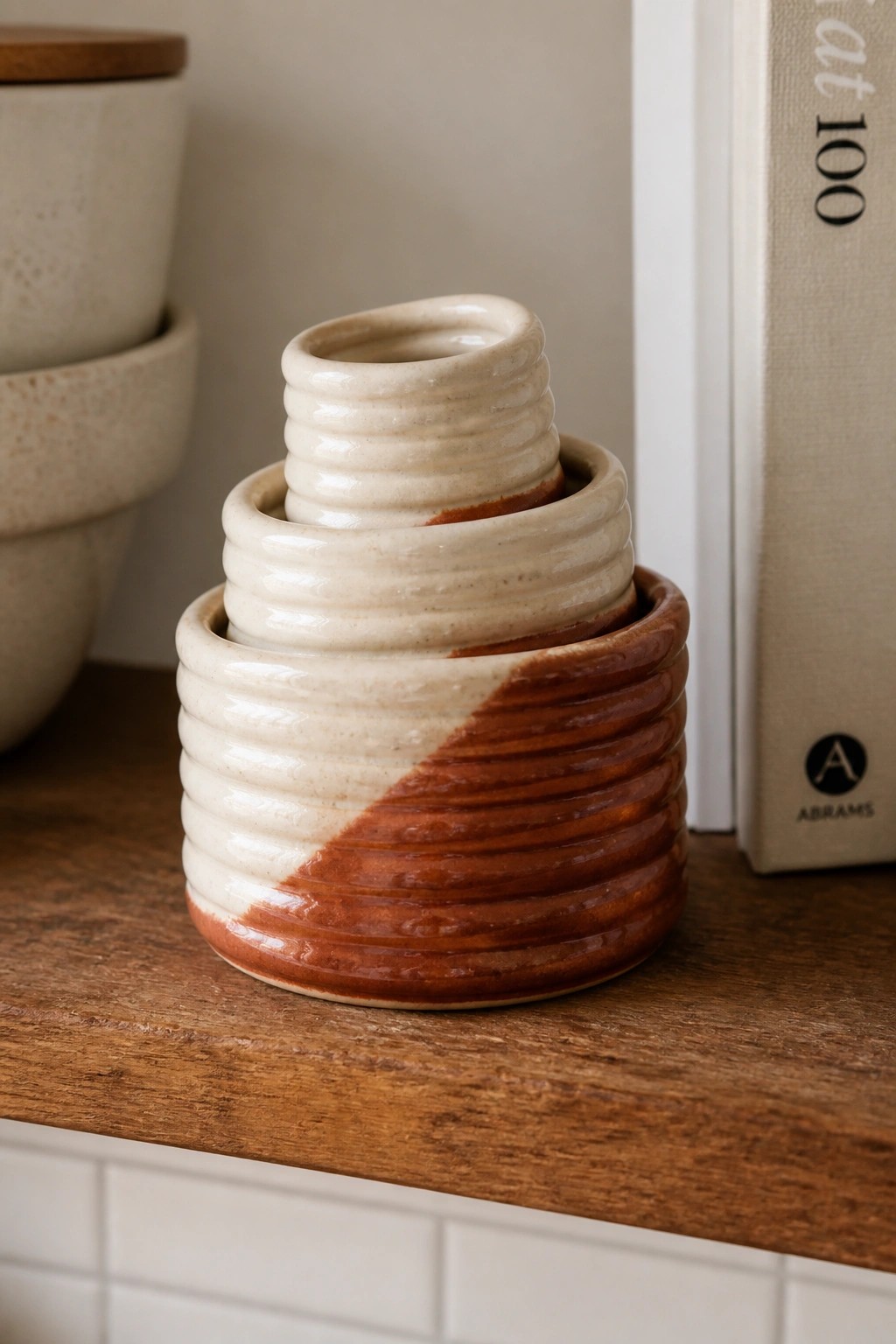 Three small ridged ceramic cylinders nested together on a wooden shelf, glazed cream on top and brown on the bottom with a diagonal divide.