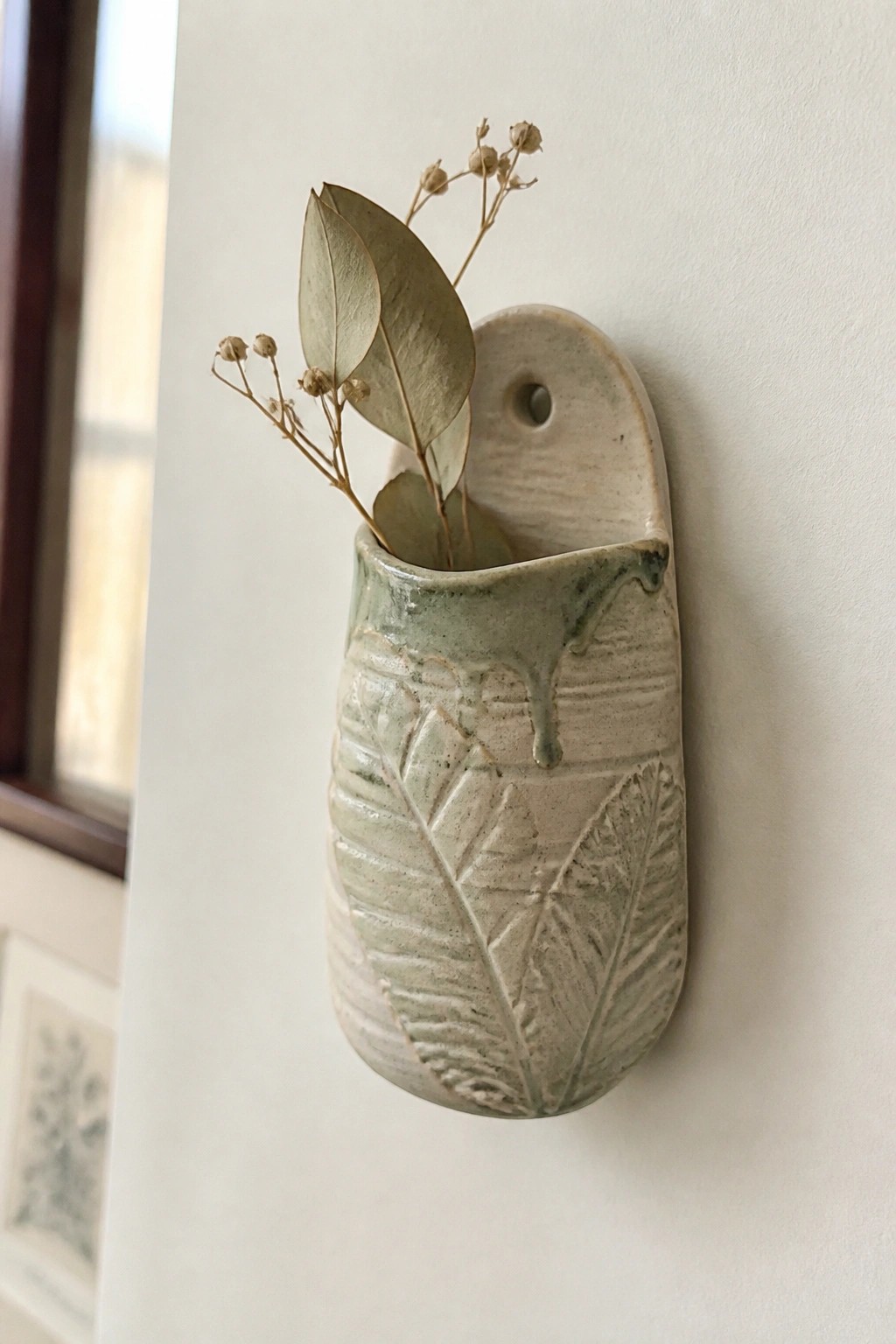 Green ceramic wall vase with embossed leaves holding dried eucalyptus stems.