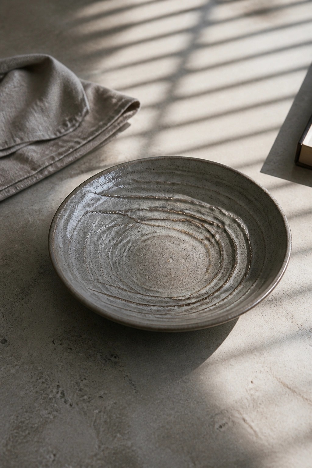 Gray ceramic bowl with concentric ridges on sunlit concrete beside linen cloth and shadows