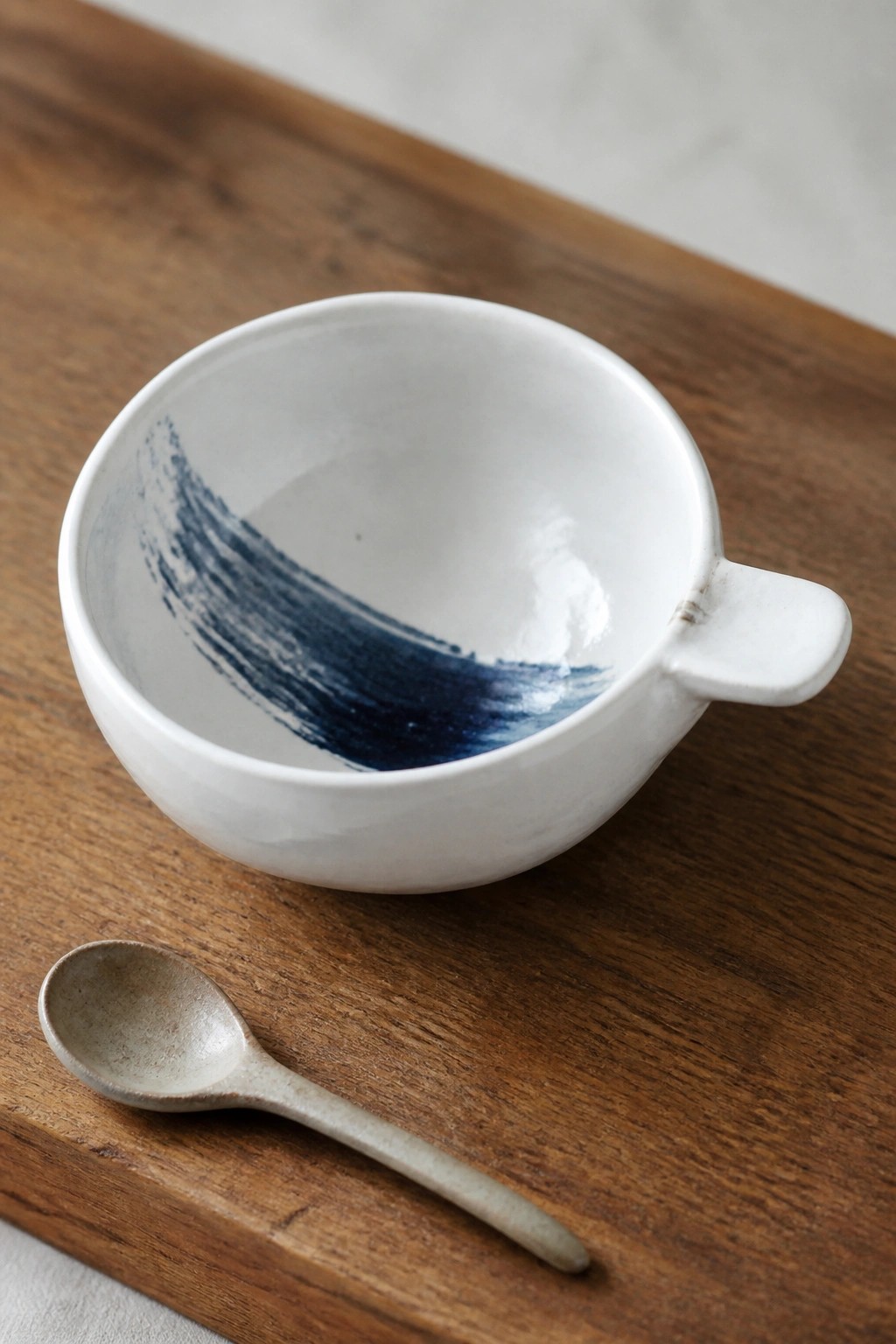 White ceramic bowl with blue brushstroke and handle beside small spoon on wood.