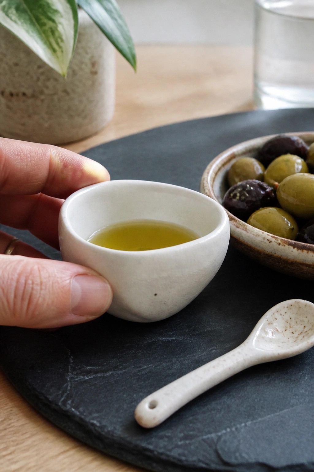 Hand holding white bowl of olive oil above spoon on slate with olives nearby