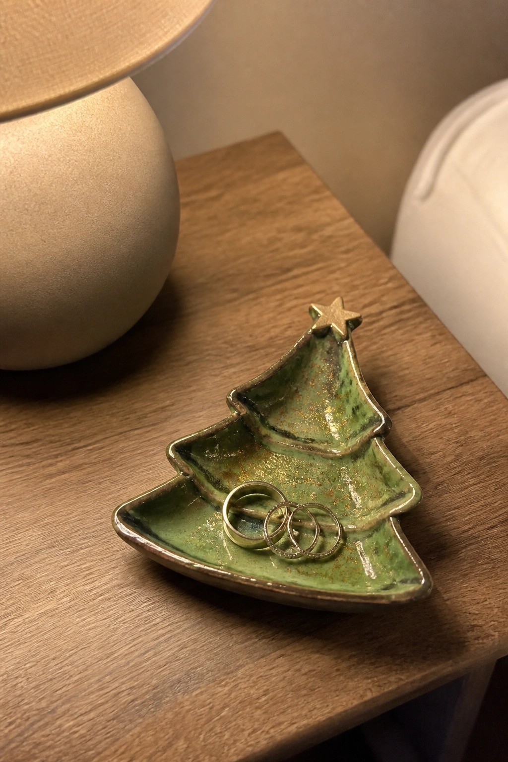 A green ceramic Christmas tree-shaped dish holding three rings sits on a wooden surface next to a lamp.