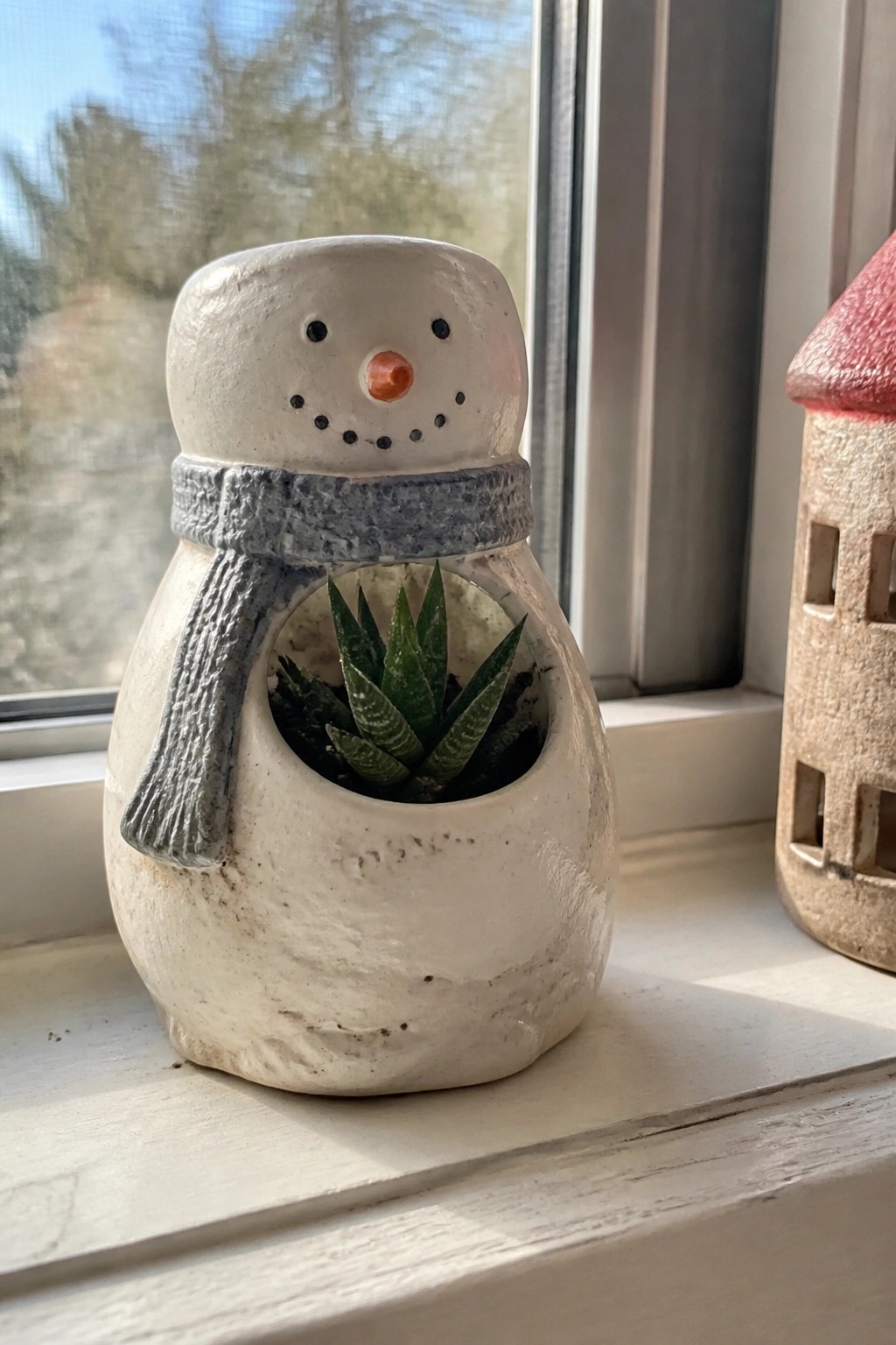 White ceramic snowman planter with gray scarf holding green succulent on windowsill.