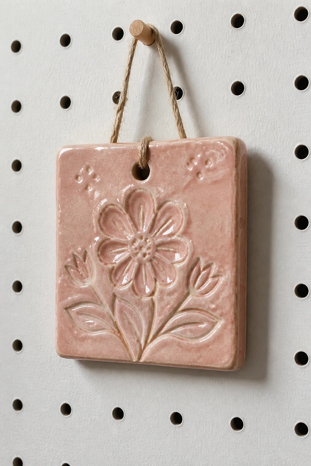 Pink ceramic tile with embossed flower and leaves, hanging by twine from peg