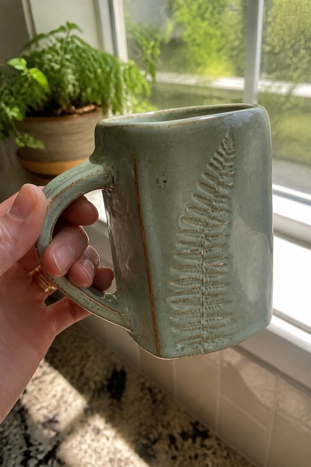 Hand with gold ring holding pale green ceramic mug with fern imprint near window