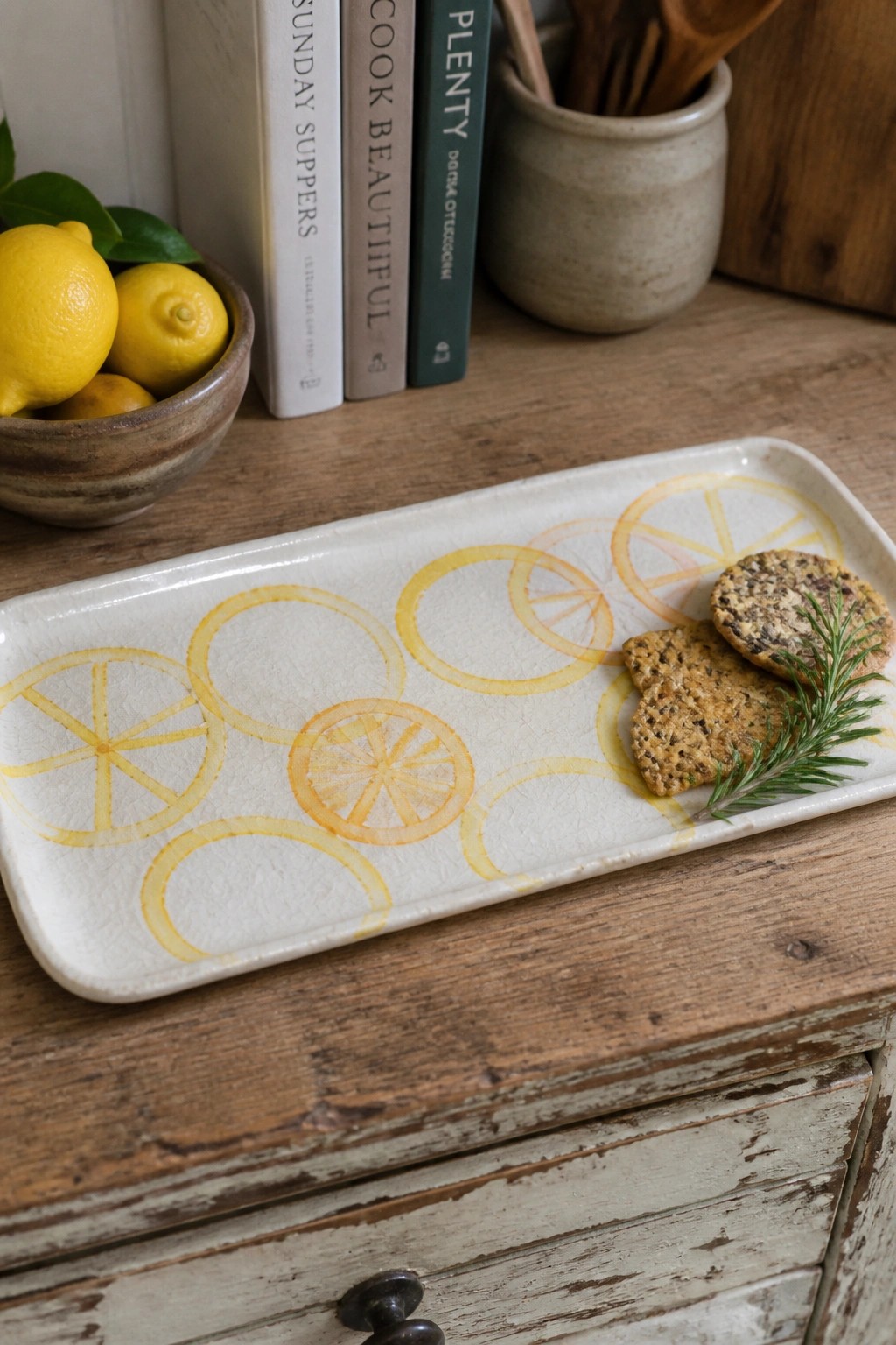 Rectangular white ceramic tray with yellow painted lemon slices holding two crackers and a rosemary sprig on a wooden surface.