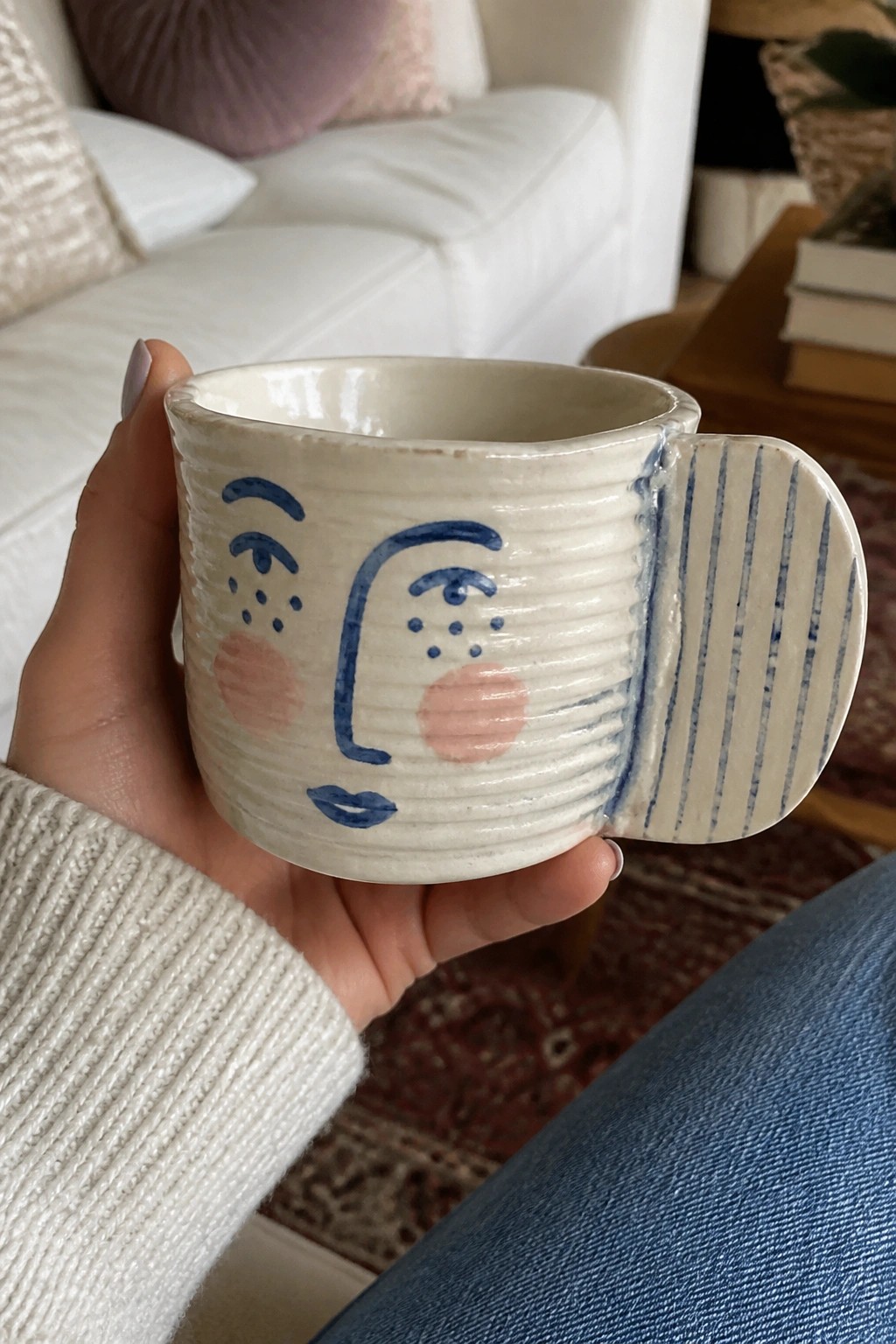Hand in white sweater holds ceramic mug with blue face and pink cheeks