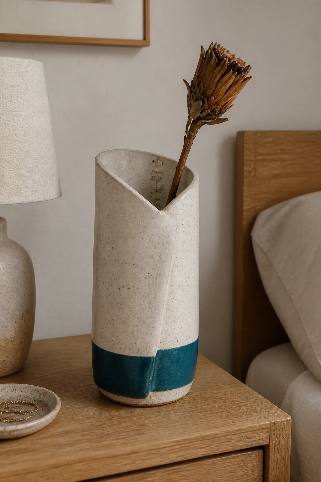 Speckled cream ceramic vase with teal band holding dried flower on wood nightstand
